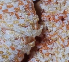 A close up of part of a ball of ribbon yarn and some fabric knitted from it. The ribbon yarn is made of three white lengthwise threads connected by many horizontal threads. Sections of matte white horizontal threads alternate with sections of glossy threads in multiple shades of orange and yellow. The spaces between the horizontal threads look very easy to accidentally stick the point of a knitting needle into.